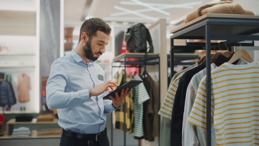Clothing Store: Male Visual Merchandising Professional Uses Tablet Computer To Create Collection. Fashionable Shop Sales Retail Manager Checks Stock. Small Business Owner Orders Stylish Items Royalty-Free Stock Footage #1071832432