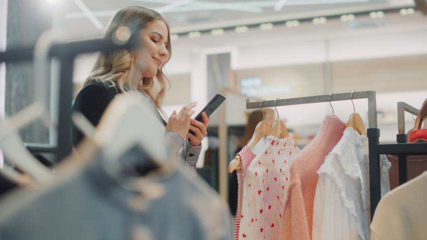 Beautiful Smiling Female Customer Shopping in Clothing Store, Using Smartphone, Browsing Online, Comparing on Internet, Choosing Stylish Clothes. Fashionable Shop, Colorful Brands, Sustainable Designs Royalty-Free Stock Footage #1071832462