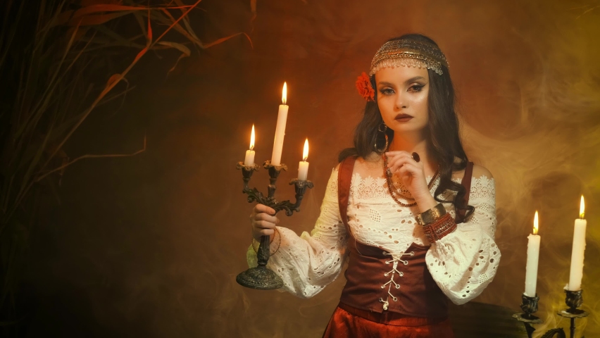 Fantasy woman gypsy in red vintage dress. Girl witch fortune teller holds candlestick burning candles. Room in smoke. Spiritual mystical seance Shows finger gesture to be silent do not make noise Royalty-Free Stock Footage #1071836017