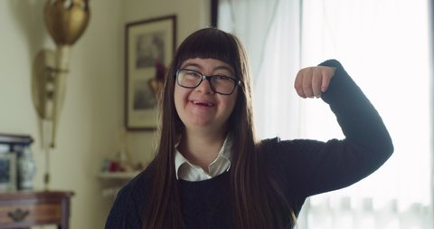 Authentic shot of happy friendly teen girl with down syndrome is showing her power with arms and smiling in camera at home. Concept of handicapped, child care, healthcare, disability, people, optimism