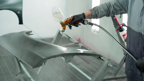 A painting technician paints a part of a car with a spray gun. The man is wearing a protective uniform. Close-up shot of hand and paint sprinkler. Slow motion