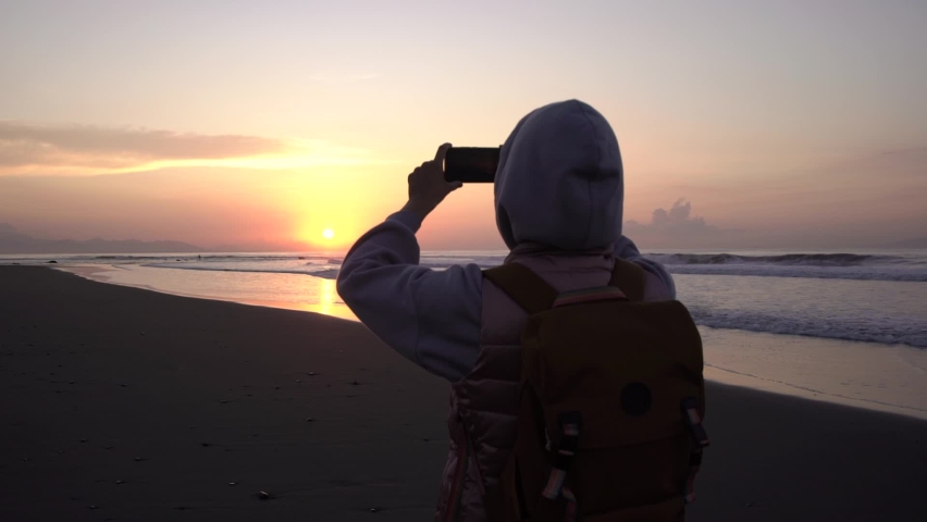 Rear view young woman traveler with backpack hood on head stands by the sea and enjoys the sunrise takes photos on your smartphone.
