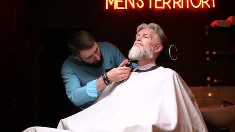 Barber work in a salon for men. A gray-haired man gets a beard trimming service
