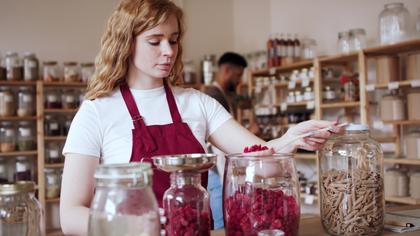 Young woman filling jar by dryed raspberries in zero waste shop. Royalty-Free Stock Footage #1071863305