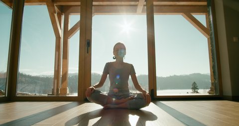 Meditating Woman in Pajamas Sits in Lotus Pose at Home with Big Window in Morning. Relax Meditation or Yoga Workout. Beautiful Shadows and Sun Lens Flare. Cinematic 4K Wide Zoom in Shot