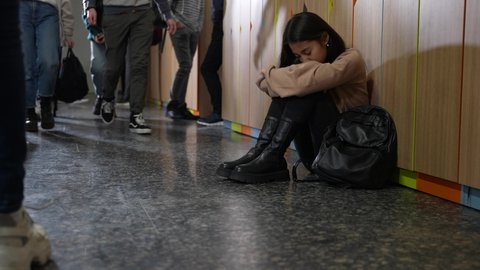 Distressed student sitting on floor by school locker while pupils walking along corridor ignoring her. Sad chinese girl sitting with head on knees while classmates leaving her out