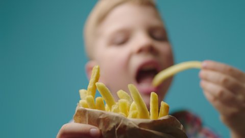 Close up of kids hand taking piece of french fries from paper bag full of fried potatoes. Boy eating french fries on blue background.