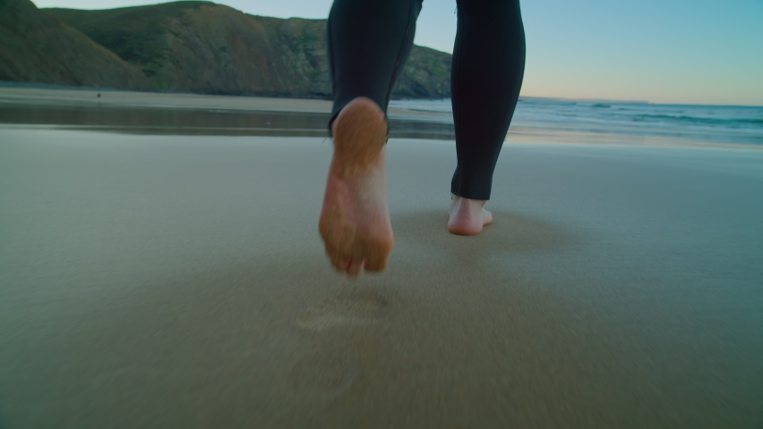 Low angle camera follow woman in wetsuit walk barefoot on wet sandy beach floor. Epic beach for surfing or cold water triathlon. Winter swimming activity Royalty-Free Stock Footage #1071884506