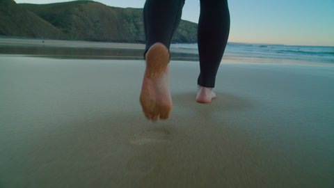 Low angle camera follow woman in wetsuit walk barefoot on wet sandy beach floor. Epic beach for surfing or cold water triathlon. Winter swimming activity