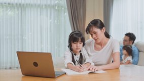 Asian happy family stay home, parents spend time with young little kid daughter in living room together. Loving mother teach and support small girl use computer for virtual learning and doing homework