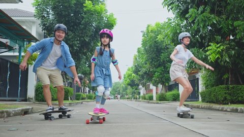 Asian active parents spend leisure time with daughter, ride Surf Skate Board on street in front of their house. Extreme sport family skater feel happy enjoy surfing together for health and well being.
