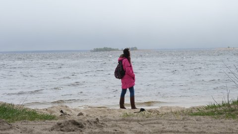 walking by the sea in winter. Sad, alone woman traveller is watching on sea or river waves, walking along sandy beach, on a windy, cold, cloudy winter day. Back view. walk along the beach in winter
