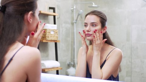Young brunette woman is applying facecream on her face in the bathroom and smiling. Morning routine: a woman in night clothes is applying makeup base on her cheeks