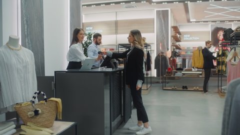 Clothing Store Checkout Cashier Counter: Beautiful Young Woman Buys Blouse from Friendly Retail Sales assistant, Paying with Contactless Credit Card. Trendy Fashion Shop with of Designer Brands