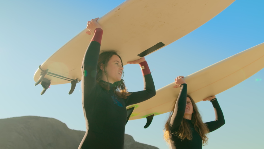 Handheld shot of two women in wetsuit outfits carry surfboards towards ocean. Cinematic sun flares camera spin around beautiful inspiring surf athletes on sunny day. Summer wanderlust vibes Royalty-Free Stock Footage #1071895261