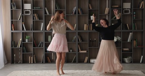 Young beautiful slim woman and her older active lively mother wear fluffy festive skirts dancing barefoot on carpet in cozy modern living room at home. Carefree family weekend, hobby and fun concept