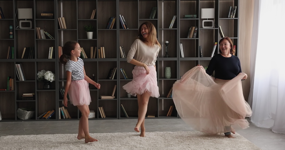 Cheerful little girl dancing in living room with young mother and 60s granny. Multi generational family diverse women wear fluffy chiffon skirts having fun on weekend at home threesome. Hobby concept Royalty-Free Stock Footage #1071895348