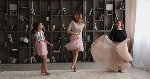 Cheerful little girl dancing in living room with young mother and 60s granny. Multi generational family diverse women wear fluffy chiffon skirts having fun on weekend at home threesome. Hobby concept