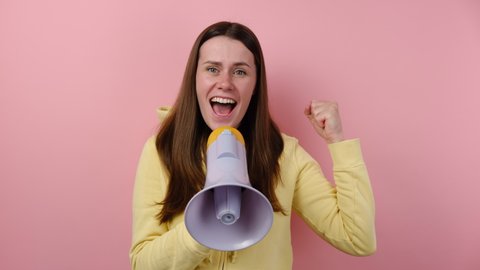 Shocked excited young woman screaming in megaphone spreading hands point finger camera on you, wears yellow hoodie, posing isolated on pink color background wall. People emotion lifestyle concept