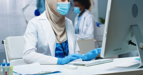 Close up of pretty young Arabic woman physician in medical mask working in cabinet in hospital typing and browsing online on computer. Covid-19 pandemic, healthcare, medical center concept