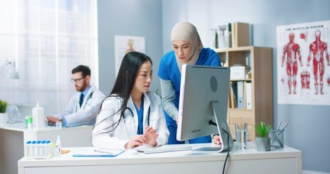 Beautiful mixed-race healthcare specialists speaking at workplace. Asian young female doctor browsing on computer and talking with Arab woman co-worker. Clinic, medic concept Video de stock