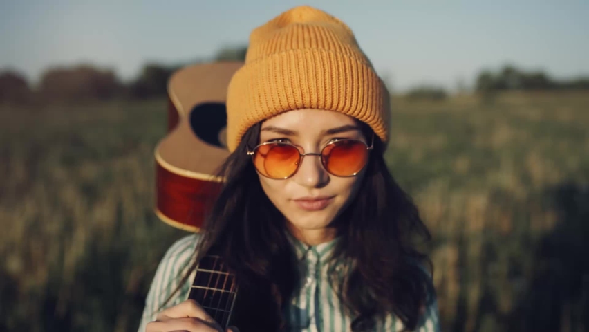 Beautiful Asian hipster woman gutar player walking on a sunny field with a acoustic. The girl in trendy sunglasses enjoy summer sunset and walking forward in countryside. Youth and creativity concept