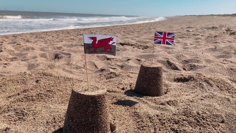 Two sandcastles on a British beach in Wales. The UK union jack and welsh flags are on top of the sand castles with the sea waves rolling in on a staycation holiday on the coast.