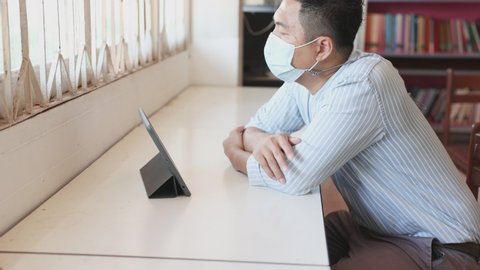 A young Asian man wears a mask to protect himself from the coronavirus infection, a young man lazily reads news about the epidemic in his home office to catch up with the epidemic in his home.