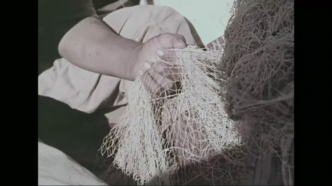 1960s: Man and boy on boat, man making fishing net. Close up of man. Close up of boy. Hand making net. Rocks in net. Boats in harbor. View of ancient ruins.