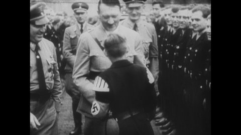 1930s: Adolf Hitler greets child in uniform at Nazi rally at Nuremberg. Hitler walks by troops standing at attention. Hitler at podium. Squadron of planes in sky. Regiment of tanks on ground.
