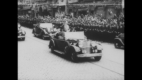 1930s: Adolf Hitler rides in convertible, giving salute, through town during Nazi rally. Soldiers march through town.