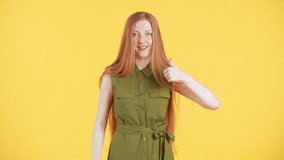 Pretty woman with freckles and long ginger hair is looking into the camera and dancing in a funny way with her thumbs up. Yellow background