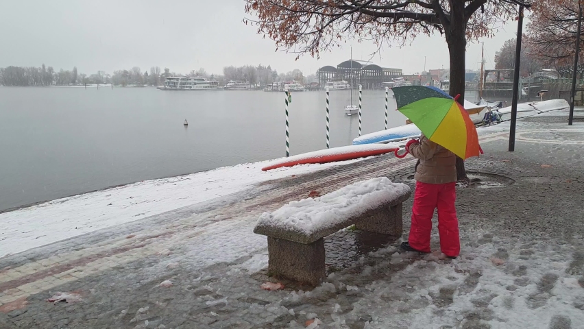 Child holding beautiful colorful umbrella plays with snow on bench in Arona. Italy