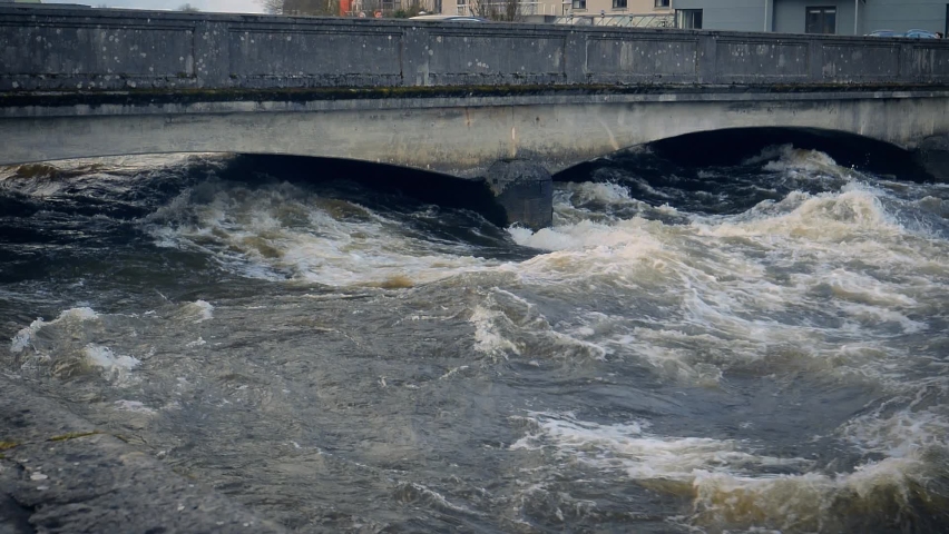 Rain swollen River Corrib rushing and churning under a bridge in Galway, Ireland. Popular Irish tourist destination and scenery of raging river under low bridge.