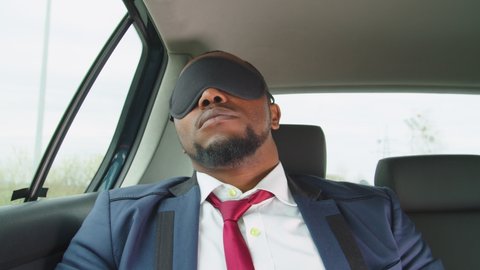 Exhausted overworked handsome african male entrepreneur in formalwear putting on sleeping mask, reclining on backseat of car to rest and nap comfortably while travelling by vehicle on business trip.