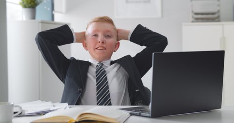 Kid businessman working on computer in office. Successful and happy little entrepreneur in formal wear relaxing in office chair working on laptop