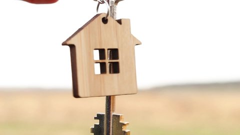 Wooden pendant of a house and key in hand. Background of the sky and field. Dream of home, building a cottage in the countryside, plan and design, delivery of the project, moving to a new house, farm.