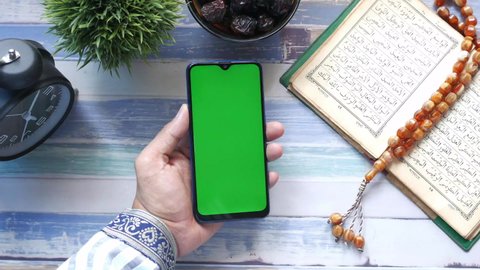 hand holding smart phone, Holy book Quran and rosary on table, 