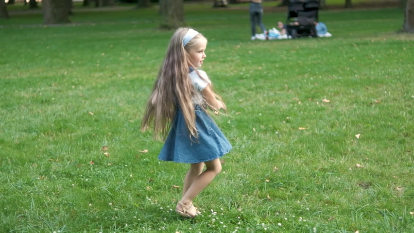 Little child girl having fun turning around on green grass in summer park.