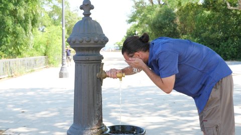 Man push lever of drinking fountain, rinse face with fresh and cool water. Guy enjoy refreshing aqua in sunny midday, hot summer weather at Barcelona.