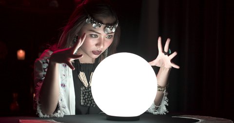Young asian woman fortuneteller performing ritual with shining crystal ball. Gypsy girl cast spell and moving hands around ball psychic for prediction. Astrology, occultism and paranormal concept.