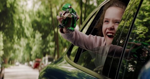 CU Portrait of cute little girl sticking head out of car window and playing with her toy dinosaur while riding through neighborhood. Shot with 2x anamorphic lens