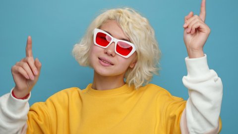 Pretty Asian Woman Smiling Dancing to Music Rhythmically to Beat Moving her Hands in Yellow Sweater Blue Background Slow Motion in Sunglasses Positive Portrait Emotions of People. Freedom. Lifestyle