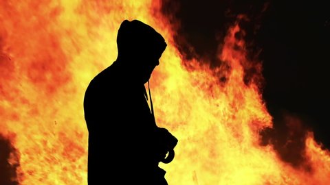 The black silhouette of a man in a hoodie, over fiery flames, unrolling adhesive tape, coming near the viewer and sealing its mouth or eyes. Symbolic shot (censorship).

