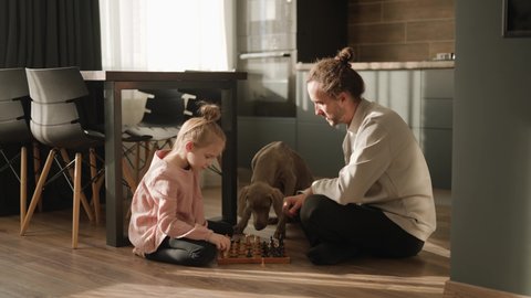 A little girl and her father are playing chess in the kitchen, sitting on the floor. A dog is standing nearby. Scandinavian and high-tech style in the interior. Family leisure