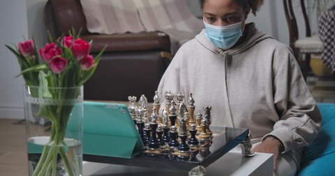 Portrait of young African American woman in Covid-19 face mask playing chess looking at tablet screen. Confident intelligent lady enjoying game online in video chat. Cinema 4k ProRes HQ