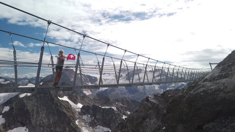 Woman with a Swiss flag on Titlis walk suspension bridge on top of Titlis mountain with glacier in the Uri Alps. Viewpoint 3028 m located in Obwalden and Bern cantons in Switzerland.
