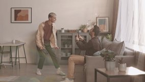 Lockdown of young Caucasian man with dark hair wearing casual clothes sitting on couch and shooting on cellphone camera his boyfriend dancing and singing in front of him