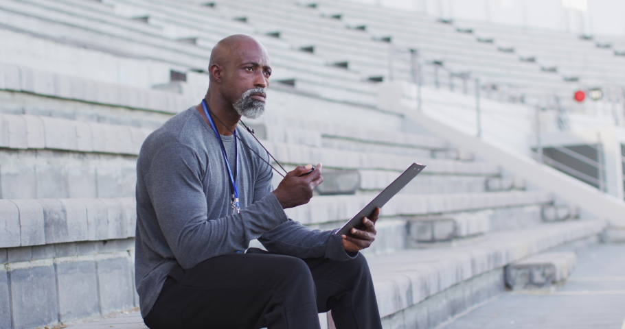 African american male coach sitting, time checking and making notes. professional runner training at sports stadium.