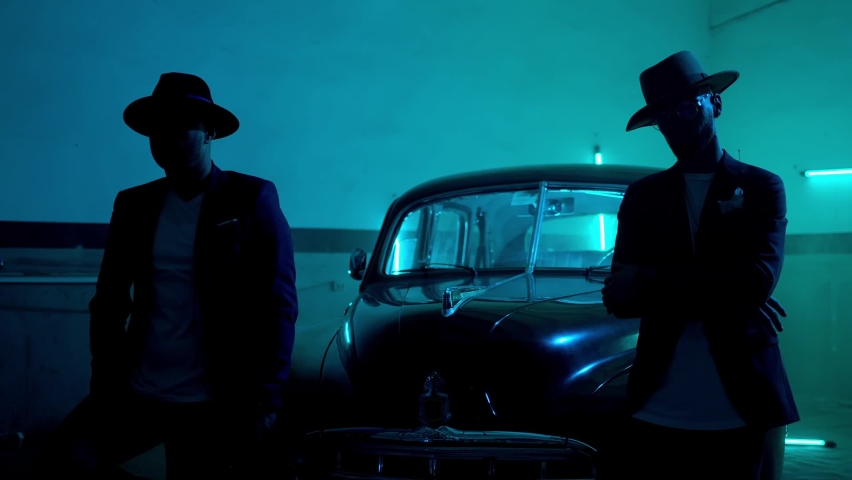 Two silhouettes of men in black suits with classic hats standing by the vintage old car and looking on camera . Dark garage interior . Strangers look like italian mafioso from movie . Slow motion shot Royalty-Free Stock Footage #1071981664